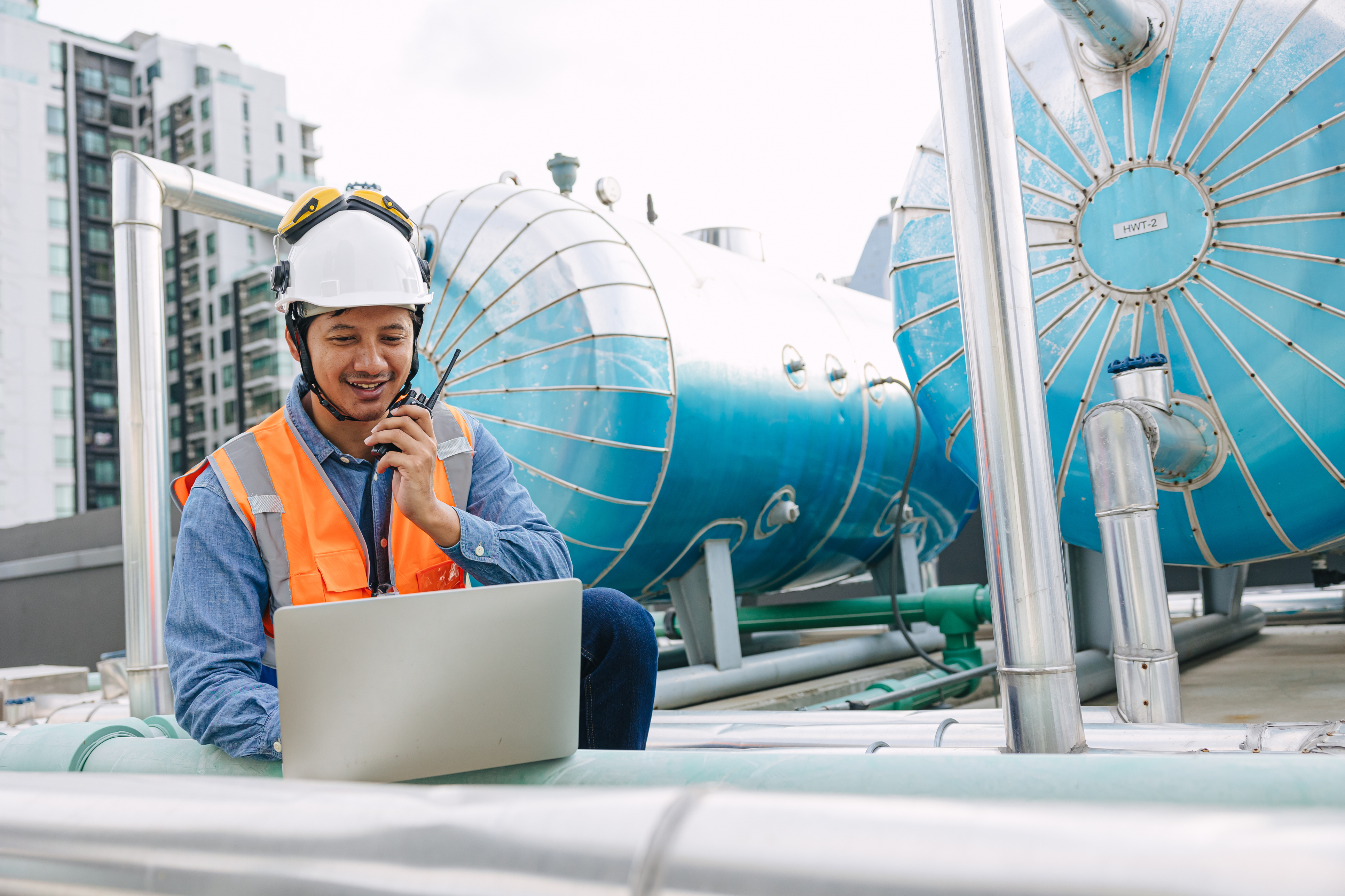 Engineer in a hard hat and safety vest using a laptop and walkie-talkie beside large industrial tanks.